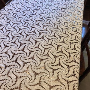May include: A white crocheted tablecloth with a swirling pattern. The tablecloth is laid out on a wooden table.