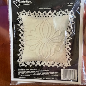 Op de afbeelding: Een witte borduurkit voor een speldenkussen, zakje of poppenkussen. De kit bevat een vierkant stuk witte stof met een bloemenmotief, omgeven door witte kanten rand. De verpakking is voorzien van de tekst "Twilling" en "Needle Magic".