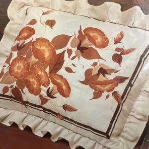 May include: A rectangular pillow with a ruffled edge featuring an embroidered floral design of rusty orange morning glories and brown leaves on a cream background. The product packaging includes the text 1816 Rusty Morning Glories (20"x 26") Crewel Kit.