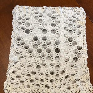 Puede incluir: Un tapete rectangular de ganchillo en color blanco roto que presenta un patrón geométrico repetitivo de pequeños cuadrados y puntos calados, terminado con un delicado borde festoneado que enmarca toda la pieza con un diseño consistente.