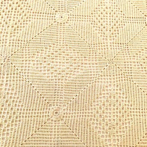 May include: A close-up of a beige crocheted tablecloth with a diamond pattern. The tablecloth has a delicate, intricate design with a repeating motif of small circles.