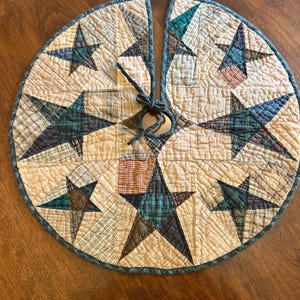 May include: A circular quilted tree skirt featuring a patchwork design of beige, tan, and cream fabric squares. The skirt is decorated with multiple five-pointed stars in various plaid patterns of blue, green, and brown. It has a center opening with ties.