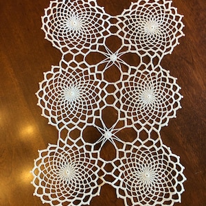 May include: A white, handmade crochet table runner. The runner features a repeating pattern of six circular doilies connected by a delicate, openwork design. The intricate lace-like details create a visually appealing and elegant aesthetic.