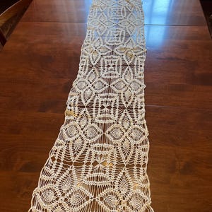 May include: Cream-colored, open-weave lace table runner. The runner features a repeating geometric pattern of circles, diamonds, and floral shapes. The delicate lace is displayed on a dark brown wooden table.