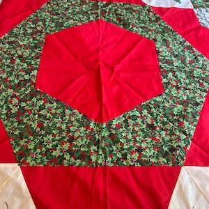 May include: A hexagonal fabric tree skirt featuring a central red panel surrounded by a green holly leaf and berry print border. The outer edge consists of alternating red and white triangular sections, with pine branch prints and white lace trim.
