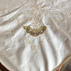 May include: Cream-colored tablecloth with intricate gold embroidery. The design features floral and leaf patterns, with a detailed, scalloped edge at the bottom. The embroidery adds a touch of elegance to the fabric.