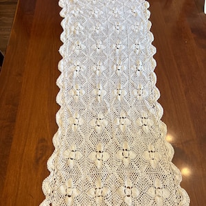 May include: A rectangular off-white crochet table runner featuring a repeating diamond pattern with central floral motifs. The runner has a scalloped edge border with a delicate openwork design throughout the entire textile piece.