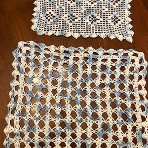 May include: Two rectangular crochet doilies featuring a white and light blue variegated yarn pattern. The top doily has a dense, intricate floral-like stitch, while the bottom doily displays an open, airy lattice design with a scalloped border.
