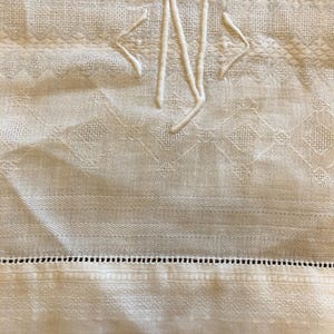 Puede incluir: Primer plano de un textil color crema con un monograma bordado. El monograma presenta las letras "N" y dos flechas estilizadas. La tela tiene un patrón texturizado y un borde decorativo con pequeños agujeros espaciados.