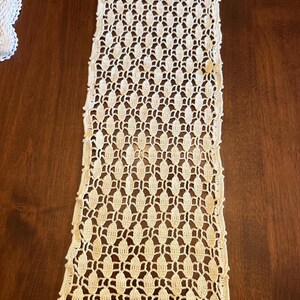 May include: A long, rectangular off-white table runner featuring an intricate, repeating crochet pattern of interconnected oval shapes. The textile has a delicate, openwork design with scalloped edges along the sides and ends, showcasing fine thread work.