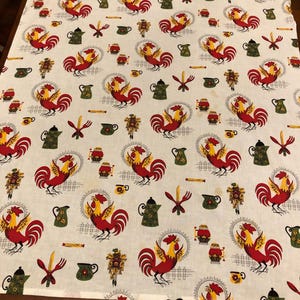 May include: White tablecloth featuring a repeating pattern of red roosters, yellow accents, green teapots, and other kitchen-themed illustrations. The rooster design is surrounded by a circular frame. The fabric is likely cotton or a similar material.