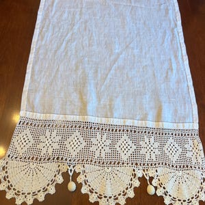May include: A rectangular white table runner made of a smooth, lightweight fabric. The runner features a wide, intricate cream-colored crochet border with a geometric floral pattern and a scalloped edge finished with small, round dangling tassels.
