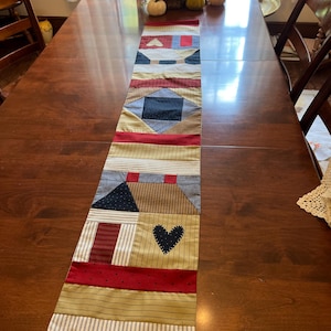May include: A long rectangular fabric table runner featuring a patchwork design with various patterns and colors including red, navy blue, tan, and cream. The runner displays geometric shapes, stripes, and a prominent dark blue heart applique.