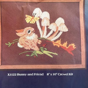 May include: An 8" x 10" crewel kit titled X1122 Bunny and Friend, featuring an embroidered scene of a tan rabbit, a yellow butterfly, white mushrooms, and autumn leaves on a brown background, framed in wood, with included materials.