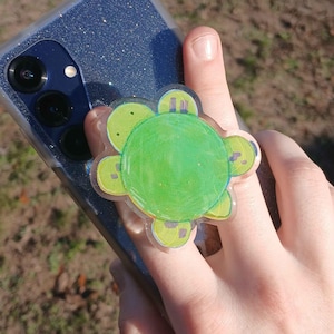 May include: A green and yellow flower-shaped phone grip is attached to a blue glitter phone case. The phone case has a camera and flash. The phone grip is held by a hand.