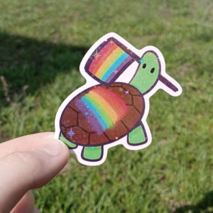 May include: A holographic sticker of a cartoon turtle with a rainbow shell and a rainbow flag. The turtle is green with a brown shell and is holding a rainbow flag. The sticker has a white border and is held up against a green background.