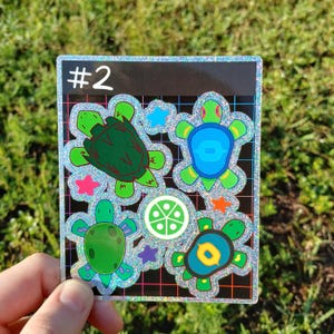 May include: A square sticker with a black background and the number "#2" in white. The sticker features four cartoon turtles in green, blue, and yellow, with star and circle accents. The sticker has a glittery, iridescent border.