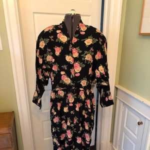 May include: A black floral dress with pink and yellow roses and green leaves. The dress has long sleeves, a button-down front, and a gathered waist. The dress is displayed on a dress form.