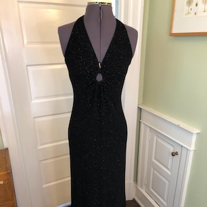 以下が含まれることがあります： 黒色の床丈ホルターネックドレス。キラキラとした質感のある生地を使用。バスト部分にはキーホールディテールがあり、体にフィットするシルエットです。マネキンに着せています。