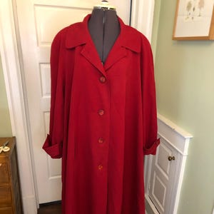 May include: A long, red coat with a classic design. The coat features a collared neckline, button-down front, and cuffed sleeves. It is displayed on a dress form with a metal stand. The coat appears to be made of a smooth, solid fabric.