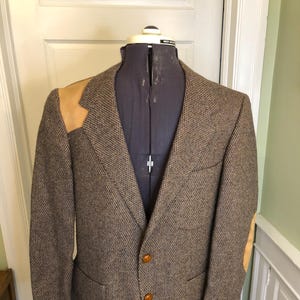 Pode incluir: Um blazer de tweed marrom com patches de camurça bege nos ombros e cotovelos. O casaco tem dois bolsos frontais, um bolso no peito e botões marrons. O blazer é exibido em um manequim.