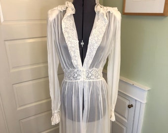 May include: A long-sleeved, floor-length white sheer robe featuring a deep V-neckline with lace trim, a fitted lace waistband with three small buttons, and ruffled lace detailing along the shoulders and cuffs, displayed on a dress form.