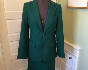 May include: A dark green skirt suit with a blazer and a skirt. The suit is on a white mannequin and is in a room with white walls and a wooden floor.