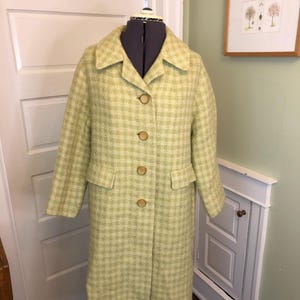 May include: A vintage, light green coat with a checkered pattern. The coat has a collared neckline, two front pockets, and three large, round buttons. The coat is displayed on a mannequin.