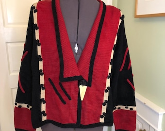 May include: A red, black, and beige knit sweater with a unique design. The sweater features a V-neck, a large beige button closure, and decorative black and beige accents. The sleeves and shoulders are black, while the front is red.