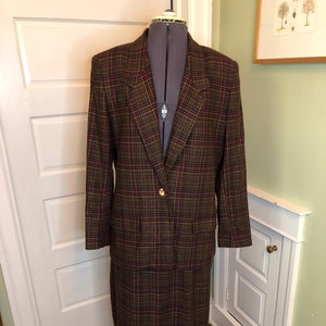 Può includere: Un completo giacca e gonna a quadri marroni, rossi e dorati. La giacca ha una chiusura con un solo bottone e una tasca sul lato sinistro. La gonna è lunga fino al ginocchio.