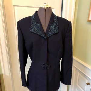 May include: A navy blue blazer with a beaded collar and a decorative clasp. The collar and clasp feature intricate beadwork in shades of teal and blue. The blazer is displayed on a mannequin, showcasing its tailored fit and classic design.