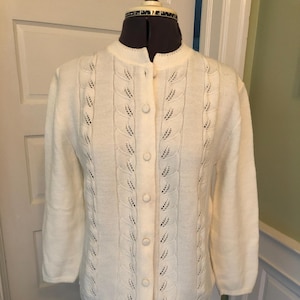 May include: A cream-coloured cardigan with a round neckline and button closure. The front panels and sleeves have a leaf-like pattern. The sleeves are three-quarter length. The garment is displayed on a mannequin.