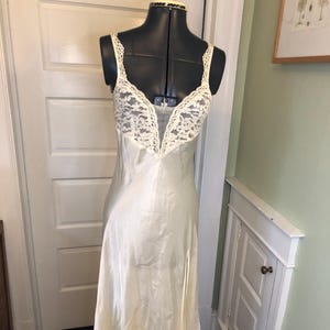 May include: Ivory-colored satin slip dress with a lace bodice and spaghetti straps. The dress has a V-neckline and a slit on the left side. The dress is displayed on a black dress form.