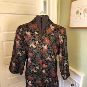 May include: A floral print blazer with three-quarter sleeves. The jacket features a black background with a pattern of red, pink, and gold flowers and green leaves. The blazer is displayed on a mannequin.