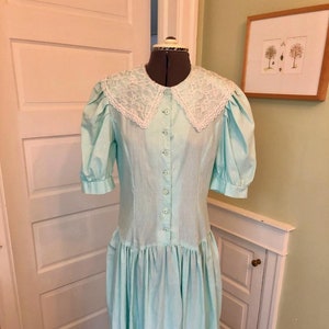 May include: A light blue dress with a white lace collar and trim. The dress has a button-up front and a gathered skirt.
