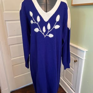 May include: A long-sleeved, knee-length sweater dress in a deep royal blue color. The garment features a V-neckline with a cream-colored trim and a matching cream-colored panel across the shoulders. A white embroidered leaf motif adorns the chest.