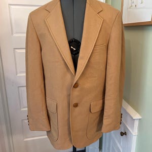 May include: A tan blazer featuring a notched lapel, two brown buttons on the front closure, and two lower flap pockets. The sleeves are finished with four small brown buttons at each cuff, and the garment is displayed on a black dress form.