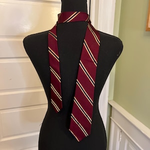 May include: A burgundy tie with gold and navy blue stripes is displayed on a black mannequin. The tie is made of a smooth fabric and is knotted around the mannequin's neck, showcasing its design and pattern.