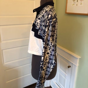 May include: A white and blue patterned cropped shirt with a black collar and long sleeves. The shirt is on a mannequin.