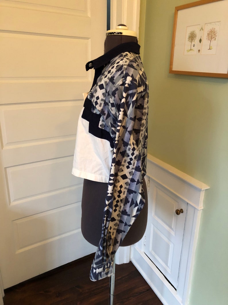 May include: A white and blue patterned cropped shirt with a black collar and long sleeves. The shirt is on a mannequin.