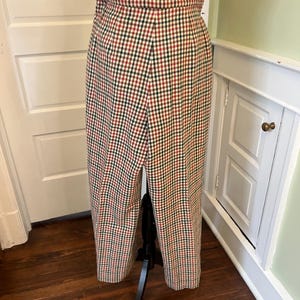 May include: High-waisted, wide-leg trousers in a classic red, green, and cream plaid pattern. The pants are displayed on a black mannequin, showcasing the pleated waist and full length. The fabric appears to be a woven material.