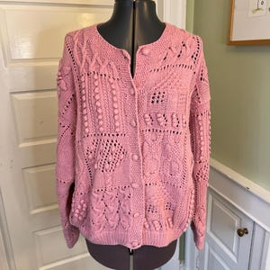 May include: A light pink, button-down cardigan sweater with a variety of textured knit patterns. The sweater is displayed on a black dress form. The knit patterns include bobbles, openwork, and cable designs. The sweater has a round neckline and long sleeves.