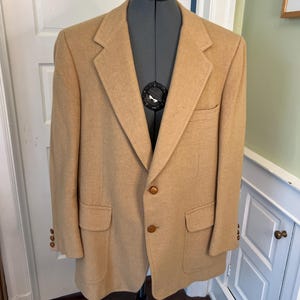 May include: A tan blazer featuring a notched lapel, two brown buttons on the front closure, two lower flap pockets, and a single chest pocket. The sleeves are finished with three small brown buttons on each cuff, and the fabric has a textured finish.