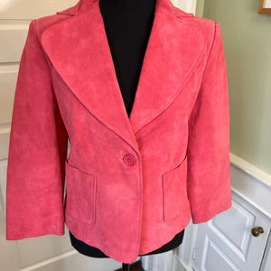 Könnte beinhalten: Korallenfarbener Wildlederblazer mit einem Knopfverschluss und zwei Vordertaschen. Die Jacke hat ein klassisches Revers und wird auf einer schwarzen Schaufensterpuppe präsentiert. Stilvolle Oberbekleidung.