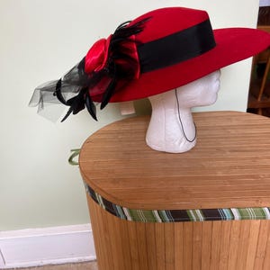 May include: A red wide-brimmed hat featuring a black satin ribbon band around the crown. The side is decorated with a red satin fabric flower, black feathers, and black netting. A thin black chin strap hangs down from the hat's interior.