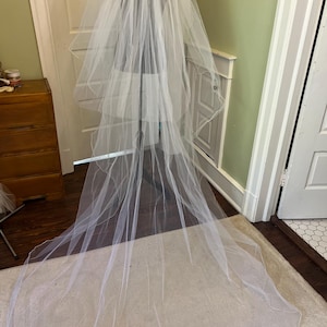 May include: A long, flowing white bridal veil, displayed on a mannequin. The veil is made of a sheer, lightweight fabric, with multiple layers cascading down. The veil is a classic wedding accessory.