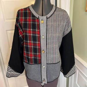 May include: A long-sleeved jacket featuring a patchwork design of red and black plaid, grey and black houndstooth, and solid black panels. The jacket has a round neckline, a front button closure with metallic buttons, and black trim detailing.