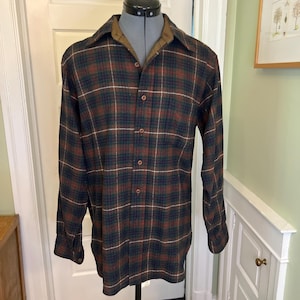Puede incluir: Una camisa de manga larga con botones y un patrón de cuadros en verde oscuro, marrón y líneas blancas finas. La camisa tiene un cuello puntiagudo, un bolsillo en el pecho en el lado izquierdo y botones marrones en la parte delantera.