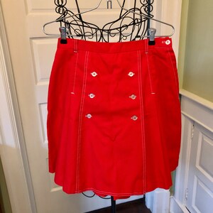 May include: A red skirt with white stitching and four silver buttons. The skirt has a slight A-line shape.
