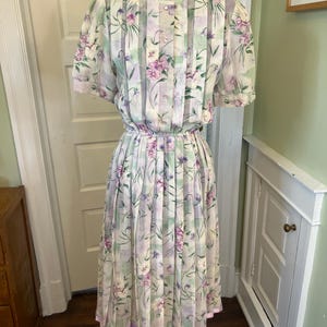Può includere: Abito floreale vintage con un motivo di fiori viola e verdi su sfondo bianco. L'abito presenta maniche corte, corpetto abbottonato e gonna a pieghe. L'orlo è rifinito con un nastro color lavanda.